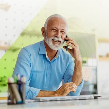 Elderly man with a white beard, smiling while talking on a phone. He wears a blue shirt and sits at a desk with stationery, conveying a joyful mood.
