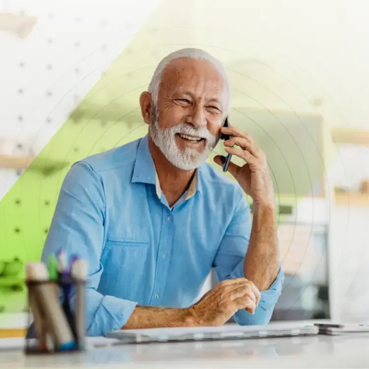Elderly man with a white beard, smiling while talking on a phone. He wears a blue shirt and sits at a desk with stationery, conveying a joyful mood.